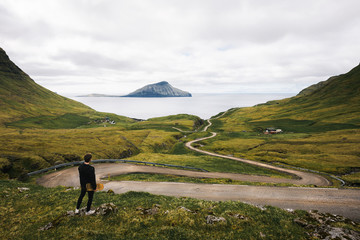 Young skater with his skateboard looks at a winding road on Faroe Islands