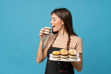 Young woman over isolated blue background holding mini cakes and eating it