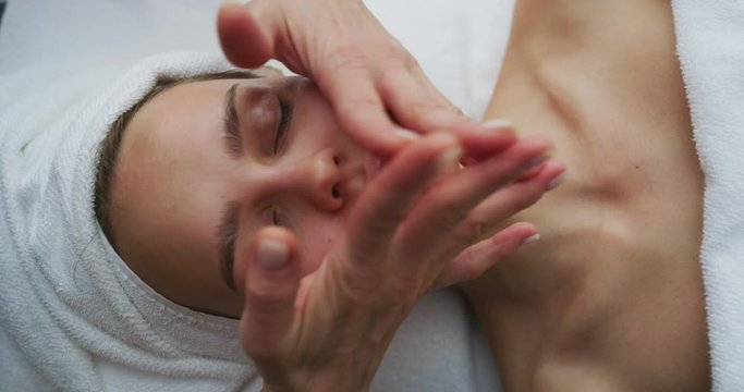 Close up of a beautiful young smiling woman with a towel on her head is receiving a facial massage and spa treatment for perfect skin in a luxury wellness center.