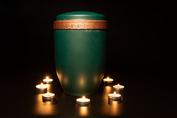 GREEN URN WITH GOLDEN EDGE, CONTAINER OF HUMAN ASHES, IN A BLACK SMOOTH BACKGROUND, surrounded with...