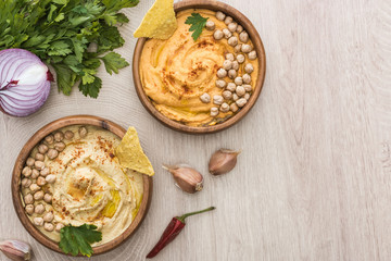 top view of delicious hummus with chickpeas, nachos in bowls near spices and parsley on beige...