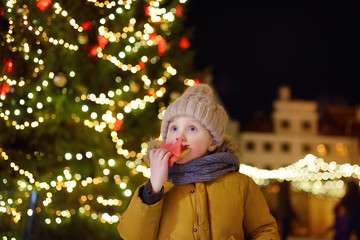 Little boy is eating snack near large Christmas fir tree. Xmas holidays on famous fair in Tallinn, Estonia.