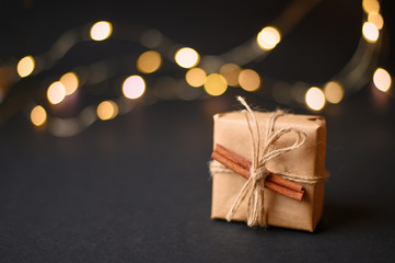 A small gift box in craft paper is decorated with cinnamon on a black photon with lights.