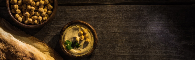 top view of delicious hummus with chickpeas in bowl near fresh baked pita on wooden rustic table, panoramic shot