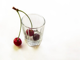 Red juicy cherry fruit with a green stalk in a glass cup. Empty transparent container with three ripe berries, close-up, isolated on a white background
