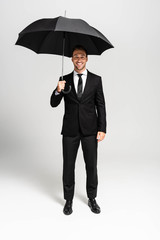 handsome and smiling businessman in suit holding umbrella on grey background