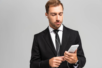 handsome businessman in suit with earphones using smartphone isolated on grey