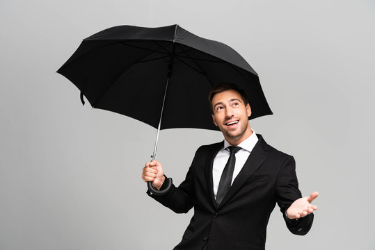 handsome and smiling businessman with outstretched hand in suit holding umbrella isolated on grey