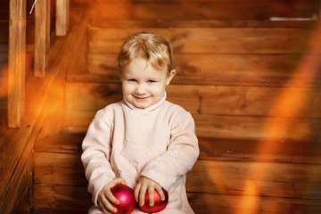 Fototapeta premium 3 years old girl in pink warm dress sits on wooden stairs and plays with red christmas balls in holiday time, winter, december. Happy and funny child in christmas lights