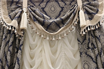 A decorative element of curtains in the design of a classic interior. Blue with beige tracks from the brushes and holders on a white Roman blinds.