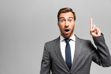 handsome and shocked businessman in suit showing idea sign isolated on grey