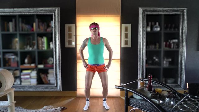 Young funny freak dressed in the style of the 80s with a mustache and glasses. On it are tight-fitting shorts and a T-shirt, the he does aerobic exercises. A meme dressing up. Guy gay. 