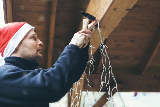 Man With Santa Hat Decorating House Outdoor Carport. Installing Christmas Lights