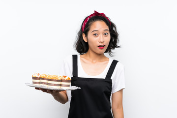 Young asian girl holding lots of muffin cake over isolated white background with surprise and shocked facial expression