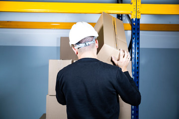 Warehouse work. Man in a hard hat placing a cardboard box on a pile. Safe work in a factory.
