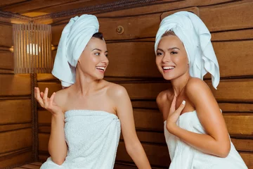 Fotobehang smiling and attractive friends in towels talking in sauna © LIGHTFIELD STUDIOS