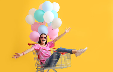 Young woman with shopping cart and balloons on color background