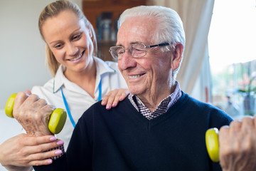 Female Physiotherapist Helping Senior Man To Lift Hand Weights At Home