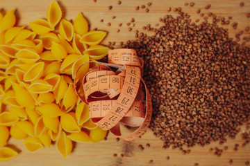 Fototapeta na wymiar Diet and weight loss theme on the orange background. Buckwheat, macaroni products and measuring tape on the wooden cutting board. Slow carbohydrates on the orange background.
