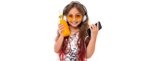 Little pretty caucasian girl listen to music with big earphones and drinks orange juice, panorama picture isolated on white background