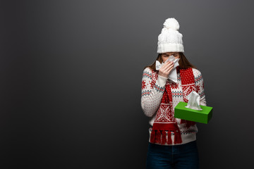 diseased woman in knitted hat sneezing with paper napkins, isolated on grey