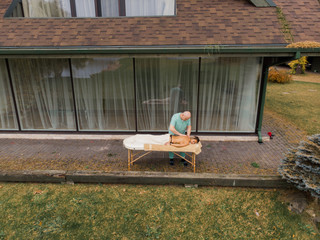 AERIAL: Blonde woman relaxing receiving back massage from male physiotherapist outdoor in wood country house lying on massage table. Young girl with tattoos relaxing destination