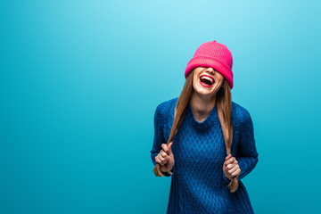 funny laughing woman in knitted sweater with pink hat on eyes, isolated on blue