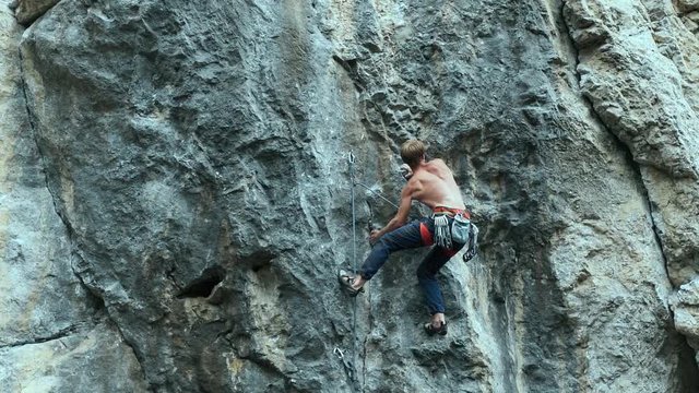 back view strong muscular man rock climber climbing on sport route outdoors on a vertical cliff, making hard wide move and gripping hold. 120 fps slow motion