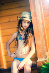 Young Girl in a swimsuit, hat and boom sitting on the railing of a bungalow on the coast - obrazy, fototapety, plakaty