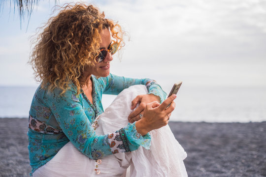 Nice cheerful caucasian woman with boho trendy clothes use modern technology cellular phone sit down at the beach in outdoor leisure activity - people with internet device cellular