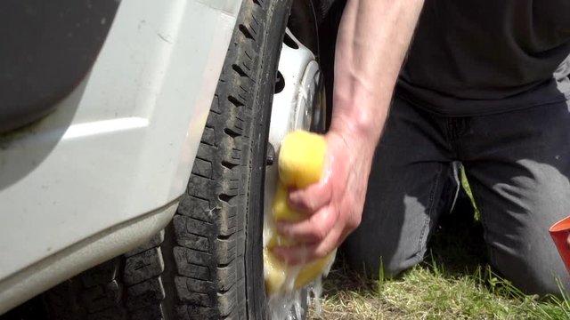 Slow motion close shot of a man’s hand using a sponge and soapy water to wash the wheel of a van.