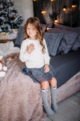 A little girl is sitting on the bed & posing on the eve of the new year 2020. Beautiful baby girl in a gray skirt and a white sweater.