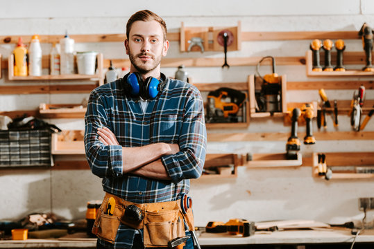 handsome carpenter in apron standing with crossed arms in workshop