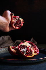 Sliced juicy pomegranate fruit on a vintage metal plate. Female hand squeezes juice from fresh pomegranate. Front view, black background, copy space