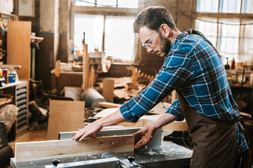 side view of bearded woodworker in apron holding plank near circular saw - obrazy, fototapety, plakaty