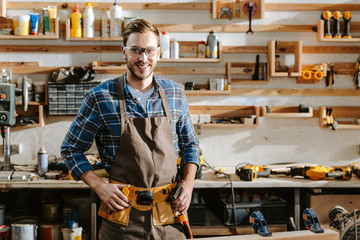 cheerful carpenter in goggles standing and touching tool belt in workshop
