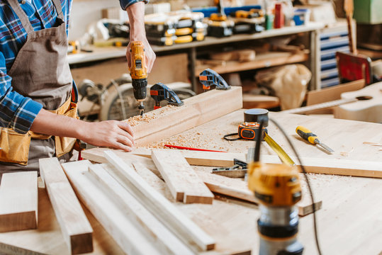 cropped view of carpenter in apron holding hammer drill near wooden planks in workshop
