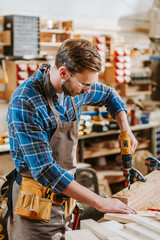 selective focus of woodworker in goggles and apron holding hammer drill near wooden planks