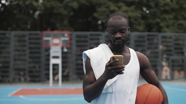 Young Muscly Serious Afro - American Guy in White Singlet Holding Ball in one hand and Using his Smartphone with other Walking at Street Basketbal Court. Healthy Lifestyle and Sport Concept.