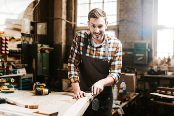 selective focus of happy bearded carpenter carving wood in workshop