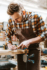 bearded carpenter in brown apron carving wood in workshop