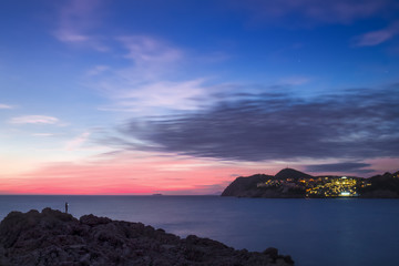 Beautiful night view to the rocky sea coast with lonely man silhouette and night city lights