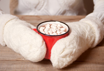Woman's hand in white mittens holding red mug of Christmas cocoa with marshmallows.