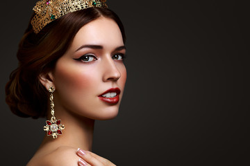 Close-up portrait beautiful girl with red lipstick in golden crown and earrings on dark background