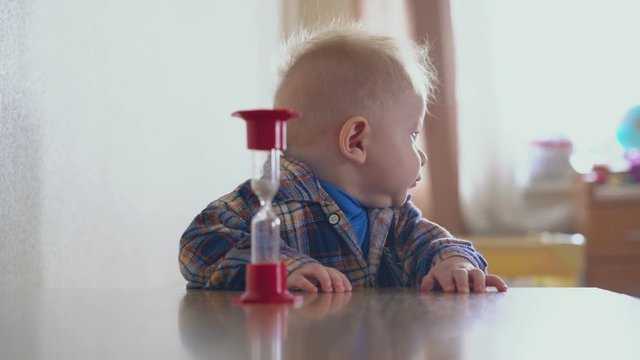 adorable funny baby in checkered shirt stands at table and mommy puts sandglass in light room close view