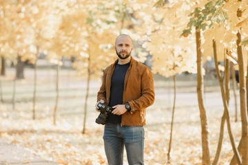 Bald brutal photographer with a beard in a suede leather jacket, blue shirt and jeans holds the camera with both hands and looks for a model in the park in the afternoon