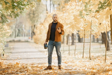 Bald stylish photographer with a beard in a suede leather jacket, blue shirt, jeans, and Chelsea boots holds the camera and looks for something in his cellphone in the park in the afternoon