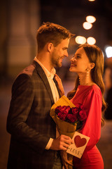 happy girl holding bouquet of roses while looking at boyfriend holding card with love inscription and card symbol