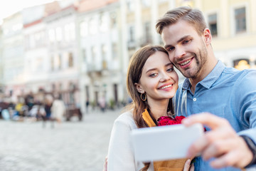 selective focus of smiling tourists taking selfie with smartphone on street