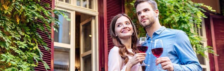 panoramic shot of happy couple holding glasses of red wine while standing near house covered with green ivy